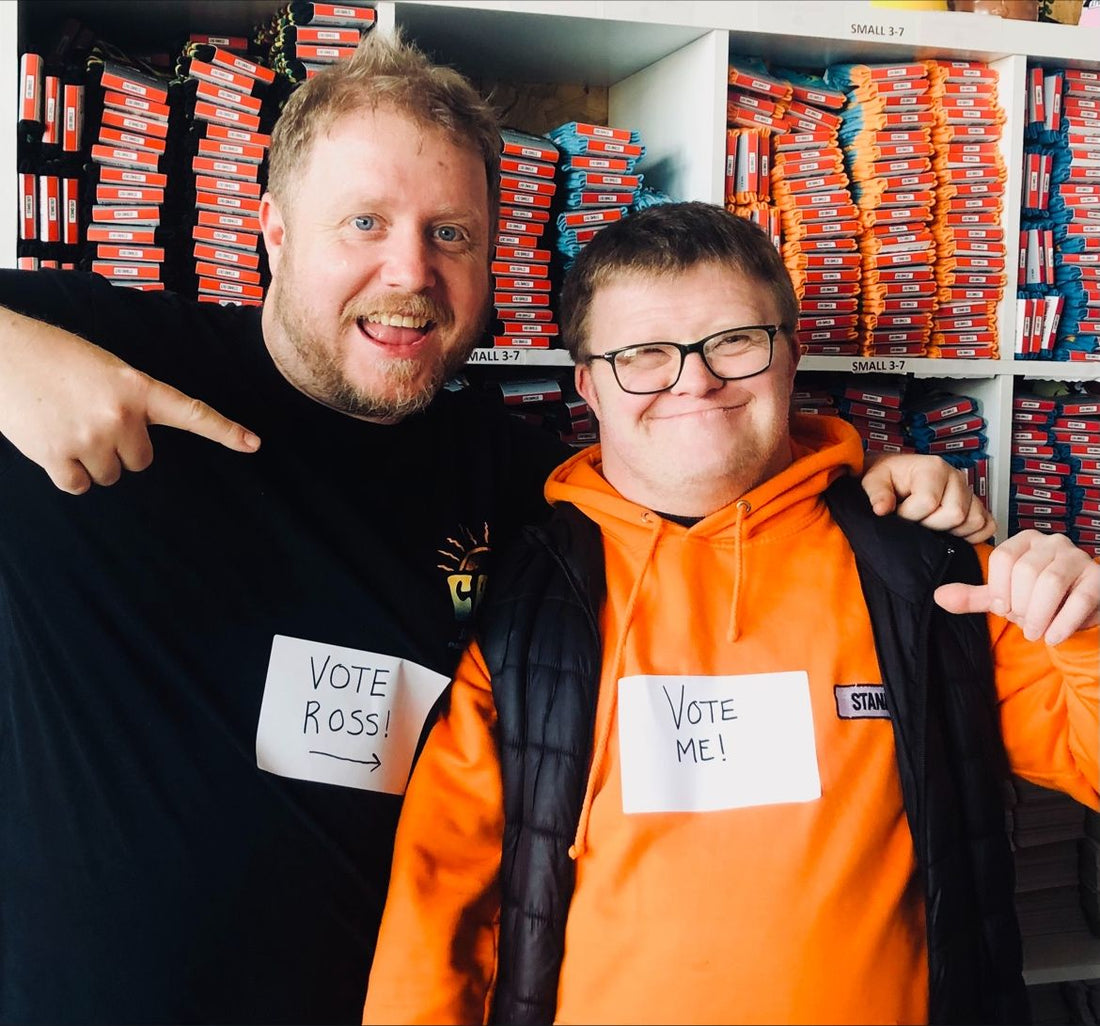 2 men standing side by side, one man who is wearing glasses has a sign on his ornage jumper saying vote me, the other man with a beard has a sign on his black tshirt saying vote ross - both men are smiling