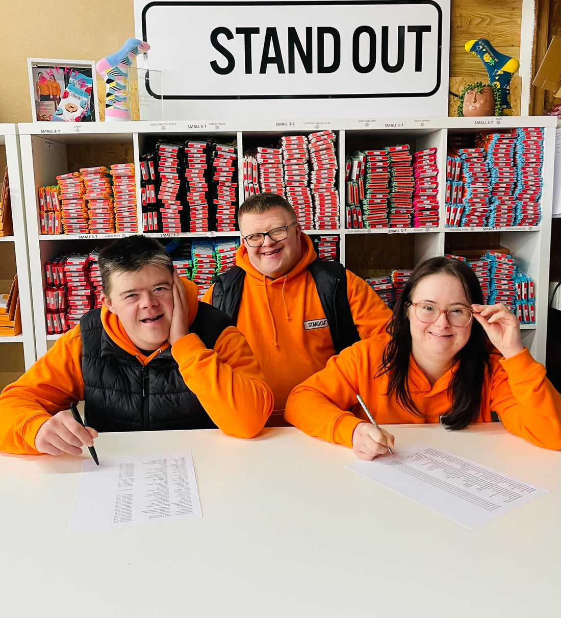 three people with orange jumpers on, a man and woman sat down and man stood behind them, all smiling