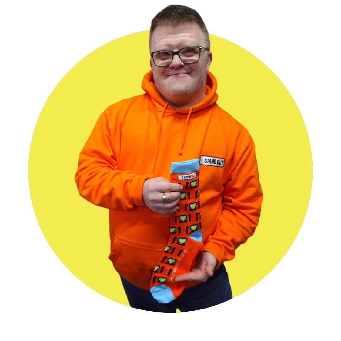 Man with Glasses on and orange jumper, Smiling and holding a pair of colourful Down Syndrome Awareness Socks