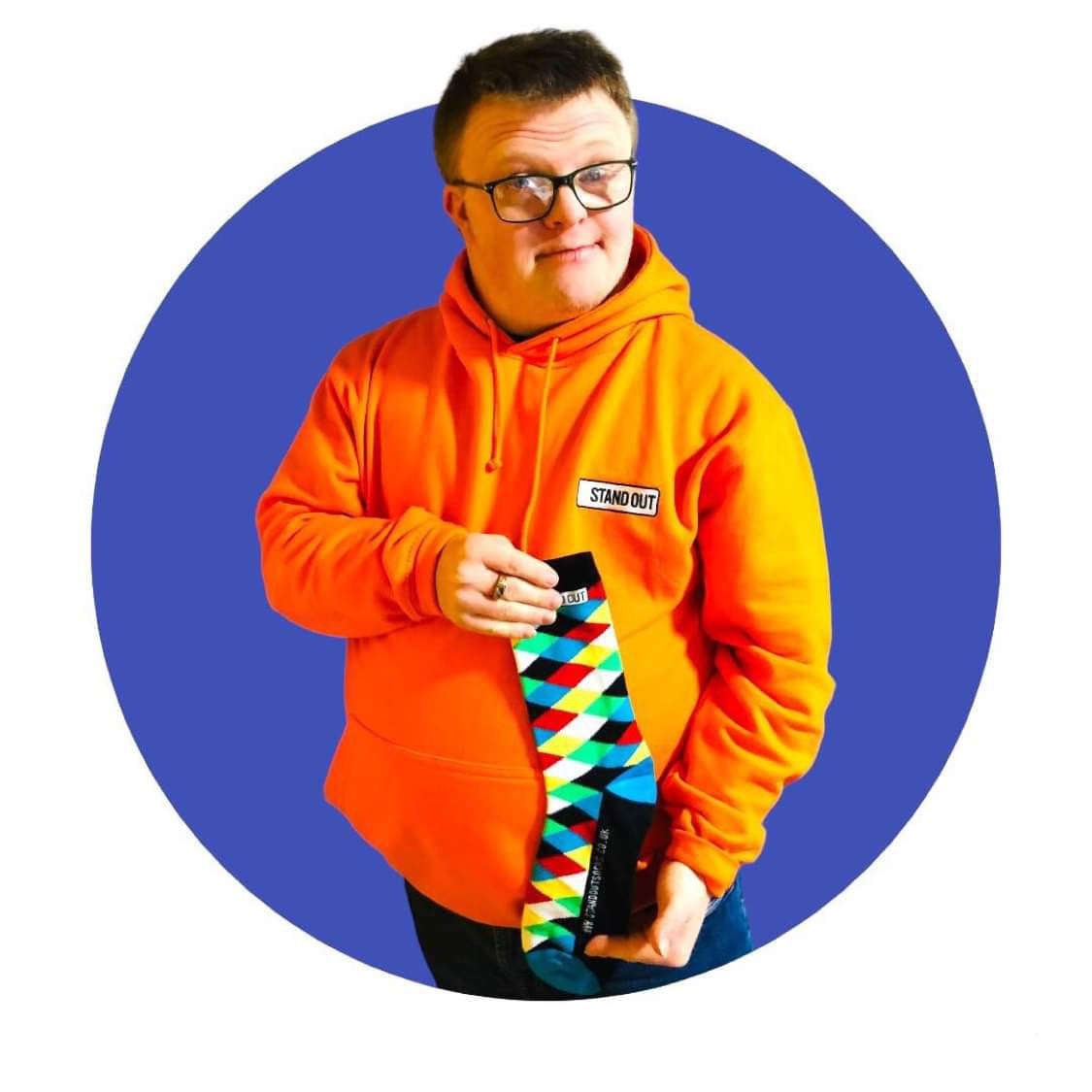 A Man wearing glasses who has down syndrome, holding up a pair of harlequin print stand out socks and he has a great big smile on his face. 