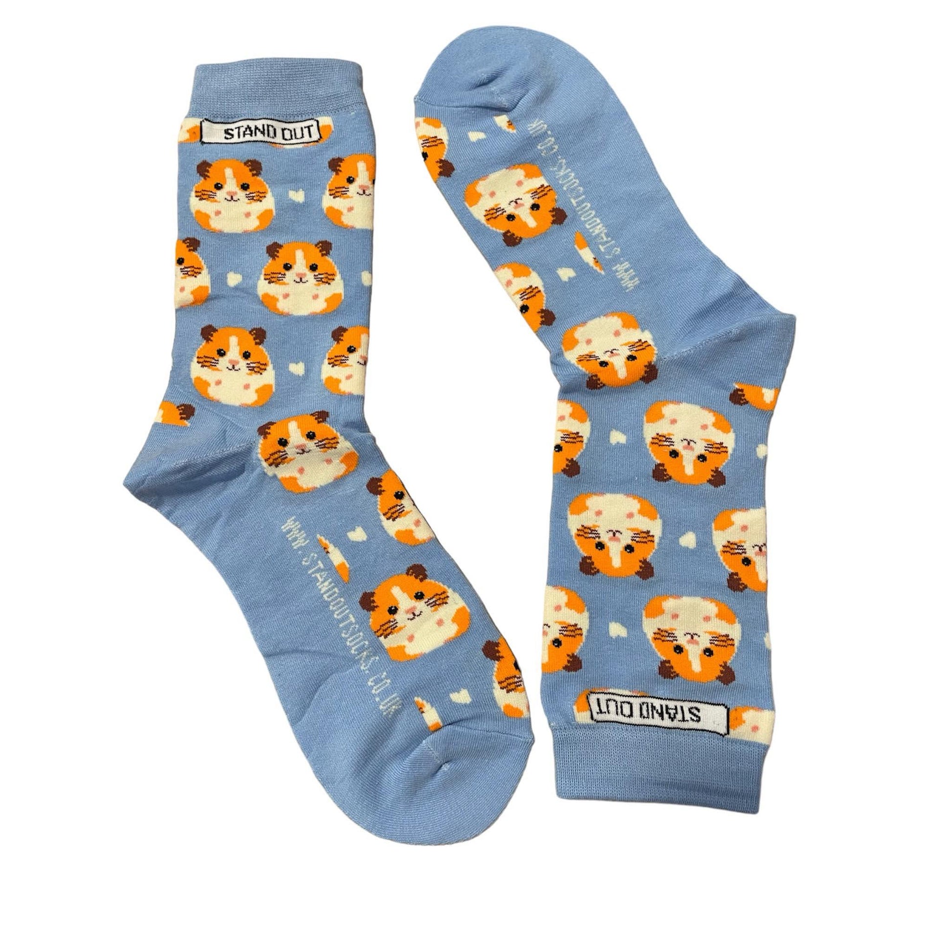 Light blue socks featuring a cute hamster pattern, created by Stand Out Socks. The socks display a playful design with orange and white guinea pig faces and small white hearts scattered throughout. The words "STAND OUT" are visible on the cuff and the company's website is printed on the sole.