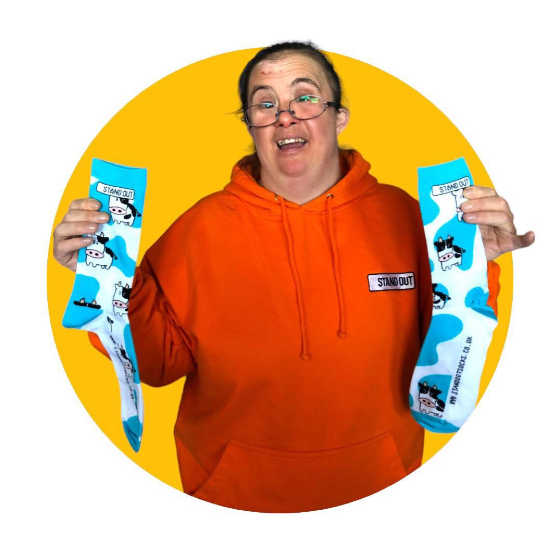 A lady who has down syndrome, wearing a orange hoodie holding a pair of blue & white cow print stand out socks 