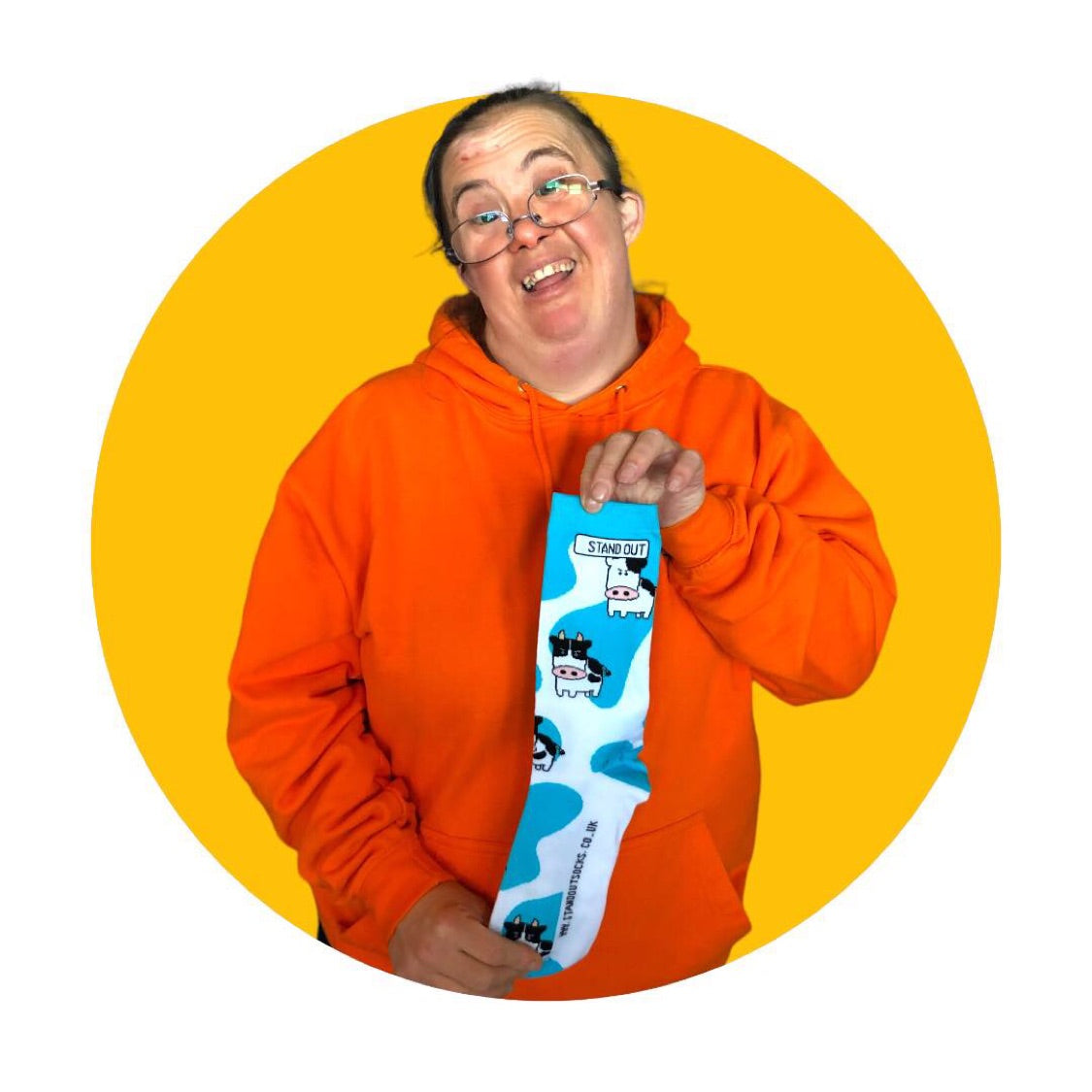 A lady who has down syndrome, wearing a orange hoodie holding a pair of blue & white cow print stand out socks