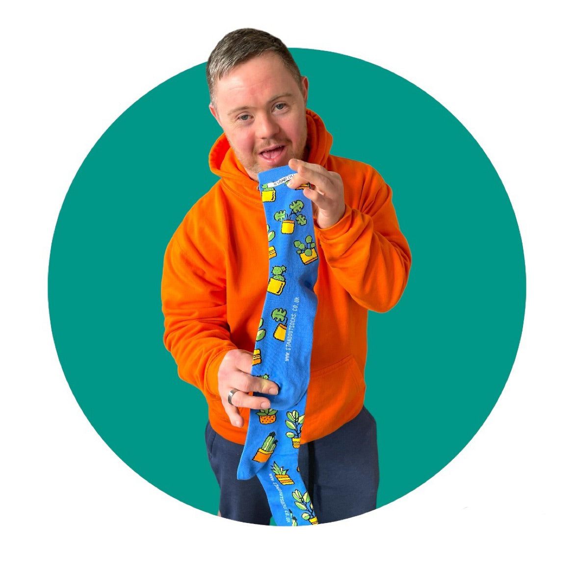 A man who has down syndrome holding up a pair of blue socks with a design of plants in pots. The picture has a green circle frame around it.,