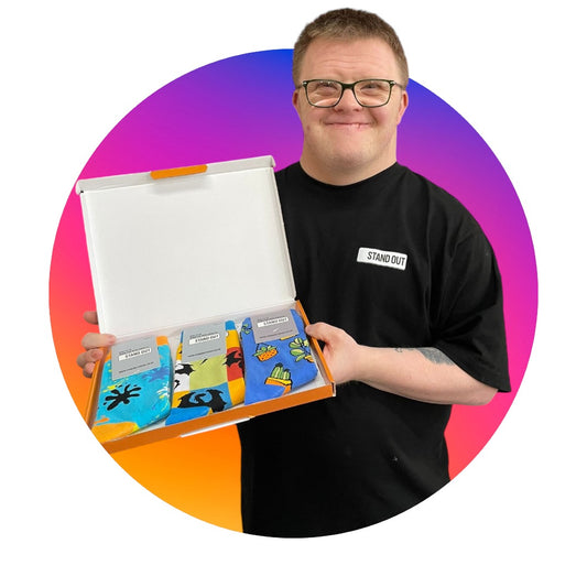 A man who has down syndrome and wearing a pair of glasses proudly holding up a box containing 3 pairs of socks made by the company ''Stand Out'' which have different funky patterns. There is a pink and purple circle frame around the man. 