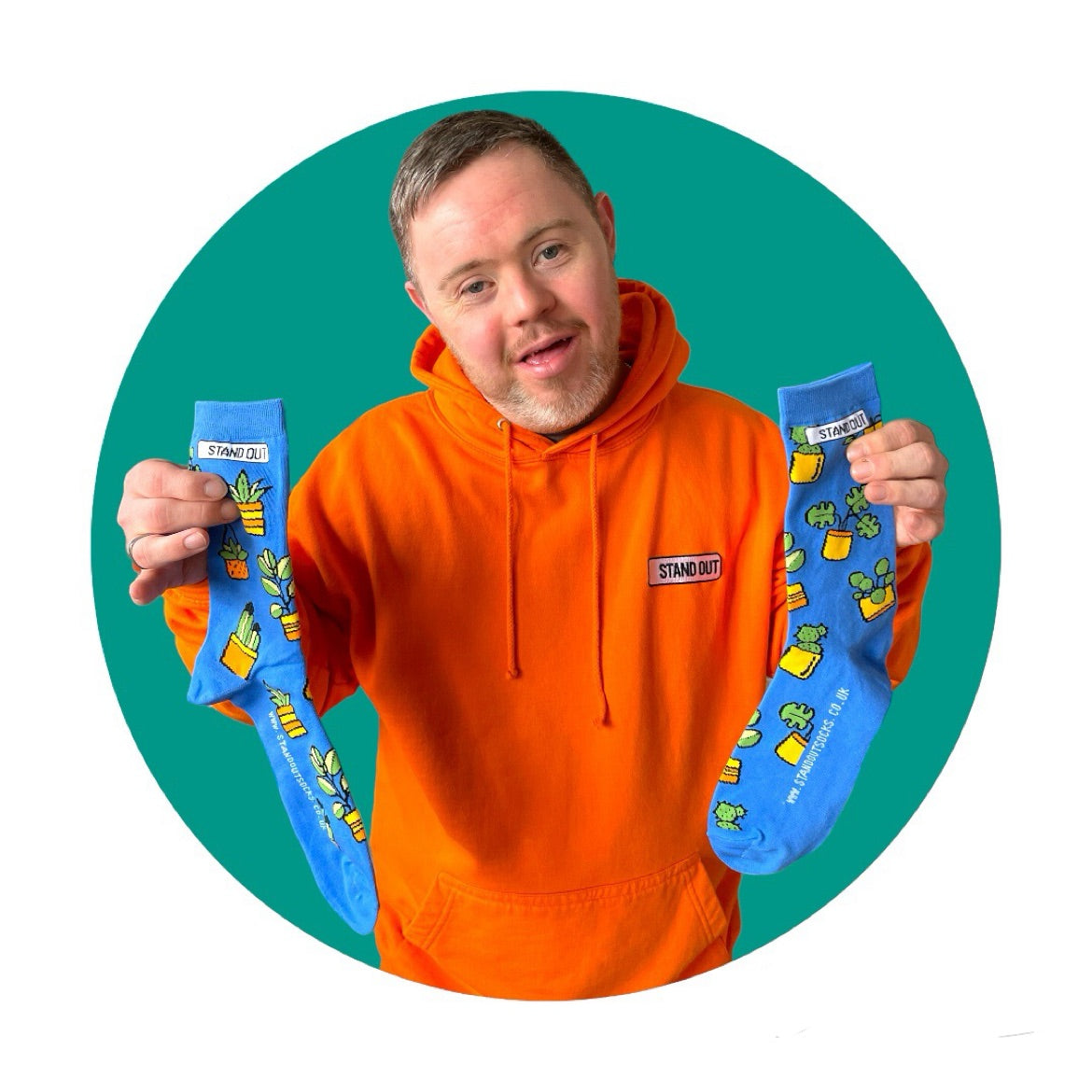 A man who has down syndrome holding up a pair of blue socks with a design of plants in pots. The picture has a green circle frame around it.,