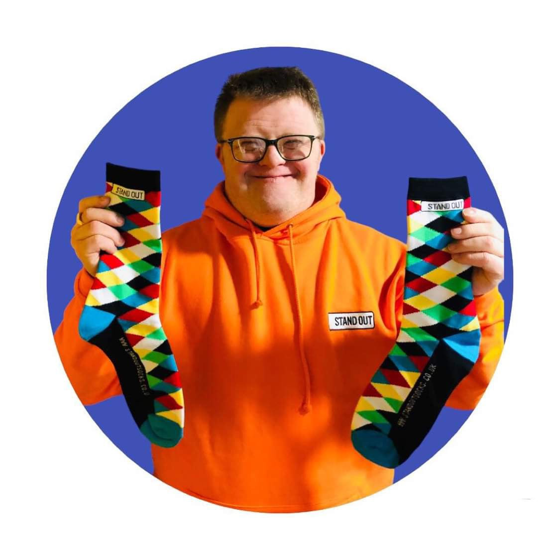 A Man wearing glasses who has down syndrome, holding up a pair of harlequin print stand out socks and he has a great big smile on his face. 