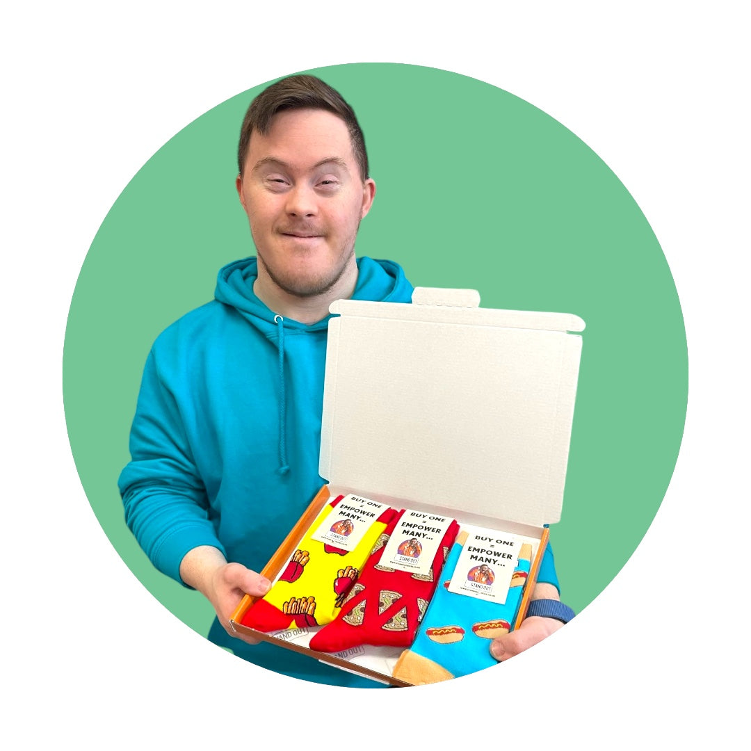 A man with Down Syndrome wearing a blue hoodie, proudly holding up a box with 3 pairs of socks with food related patterns. 