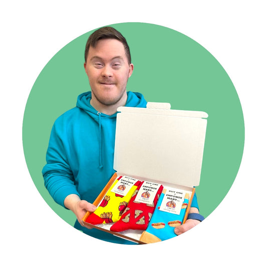 A man with Down Syndrome wearing a blue hoodie, proudly holding up a box with 3 pairs of socks with food related patterns. 