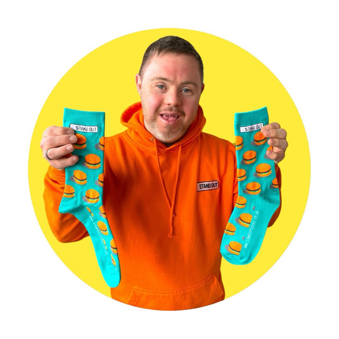 A Man who has down syndrome holding up a pair of green socks with burgers on them and smiling. with a yellow circle frame around the picture..