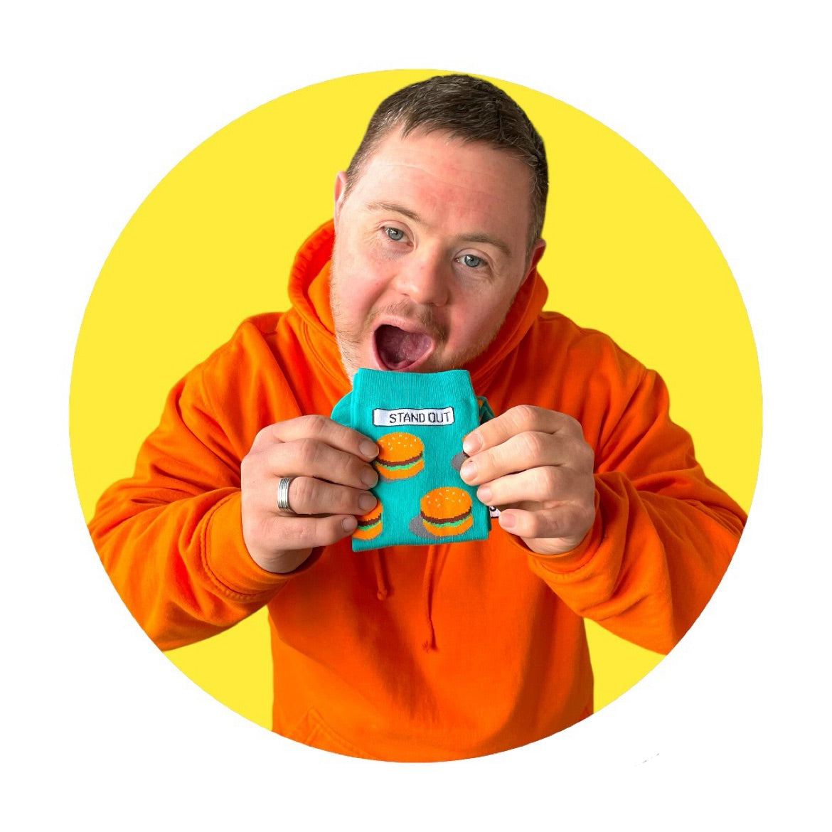 A Man who has down syndrome holding up a pair of green socks with burgers on them and smiling. with a yellow circle frame around the picture..