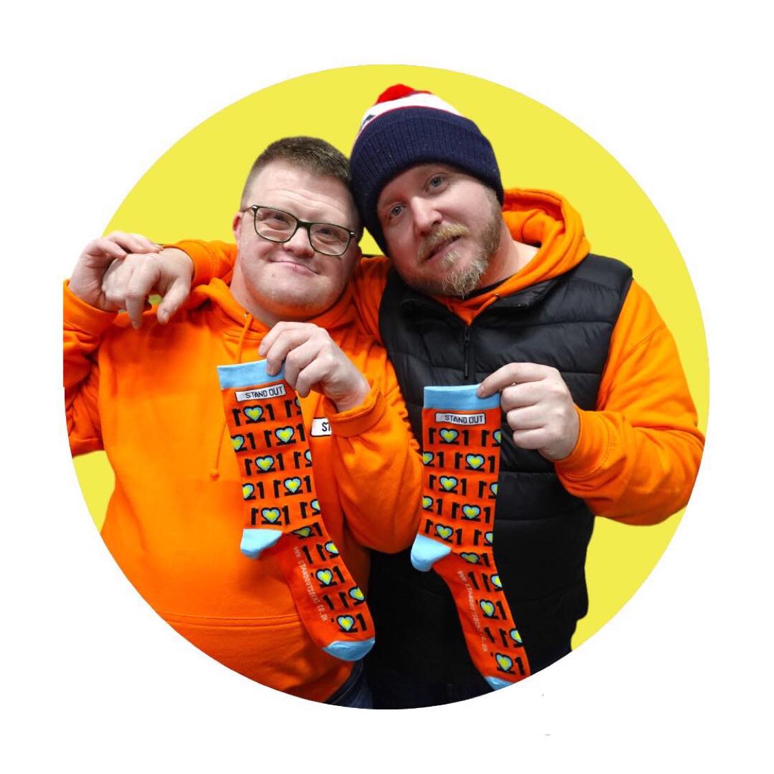 2 Men, one with  with Glasses on and orange jumper, and the other with a hat on, Smiling and holding a pair of colourful Down Syndrome Awareness Socks