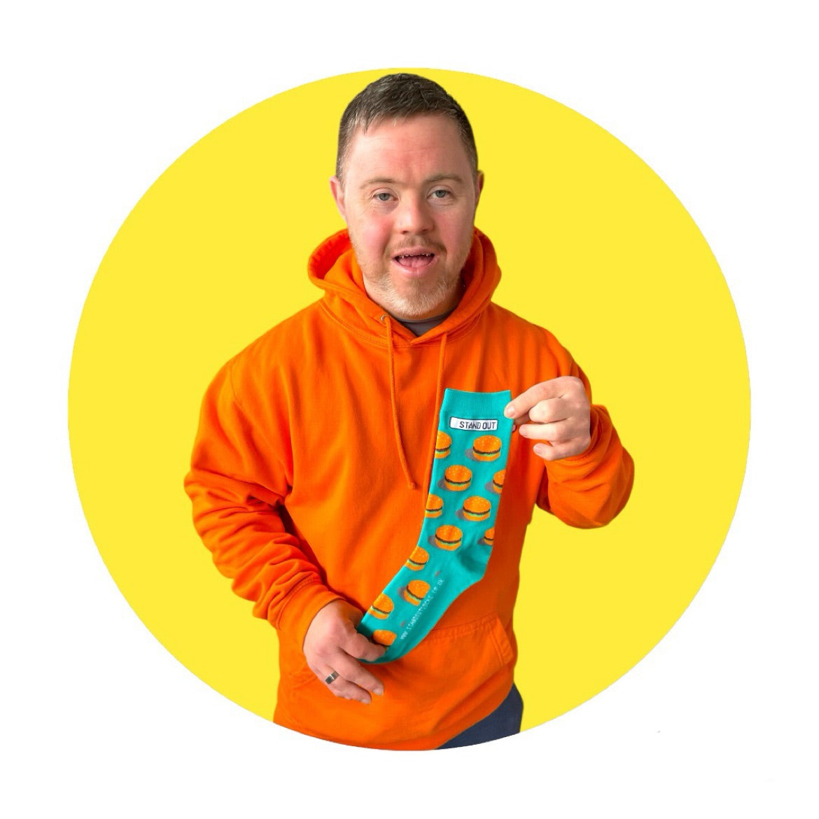 A Man who has down syndrome holding up a pair of green socks with burgers on them and smiling. with a yellow circle frame around the picture.